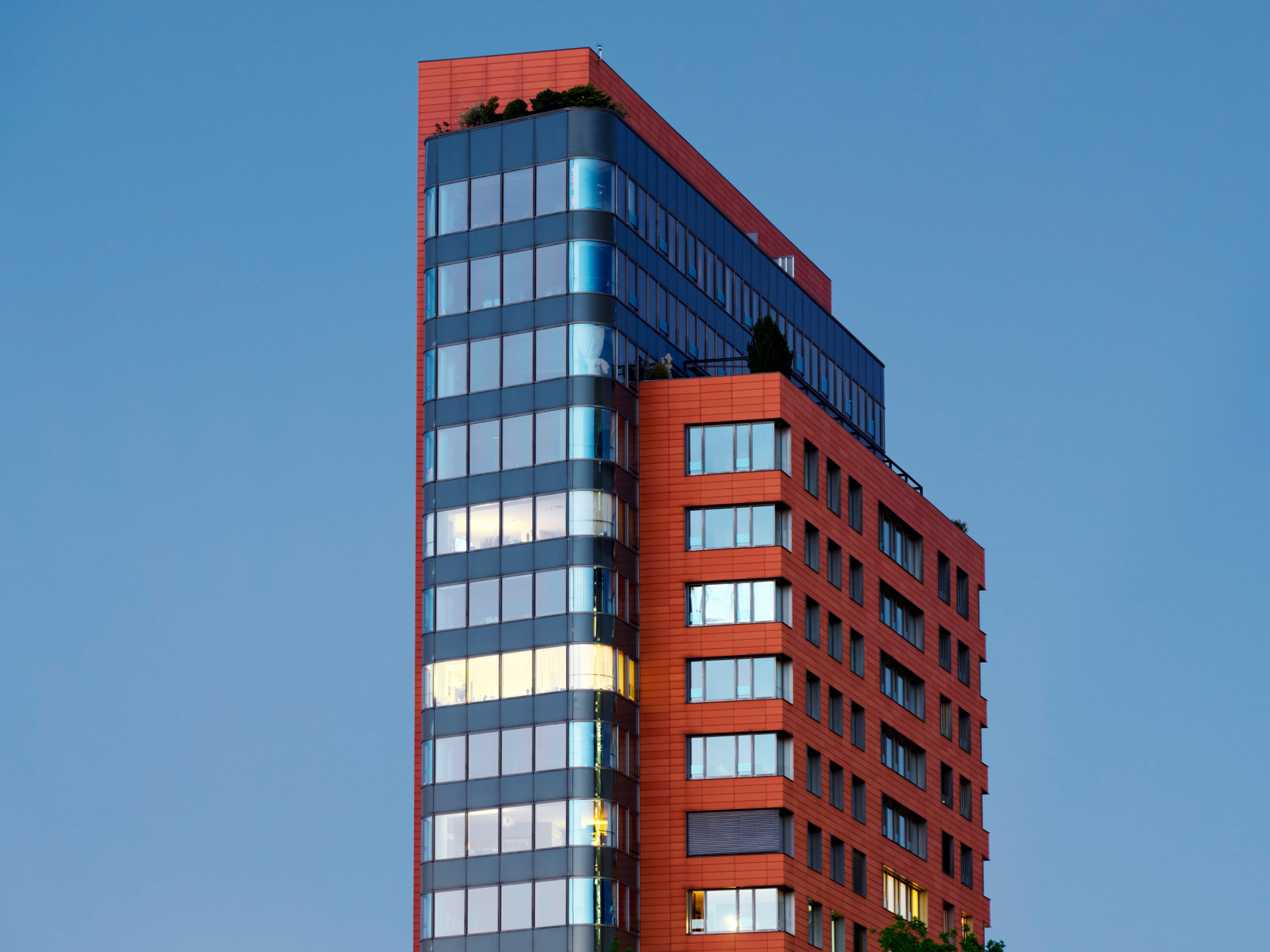 Das Portobello Haus in Düsseldorf. Ein modernes Hochhaus mit spiegelnder Glasfassade und rötlichen Paneelen, sichtbarer Dachbegrünung, bei klarem Himmel.