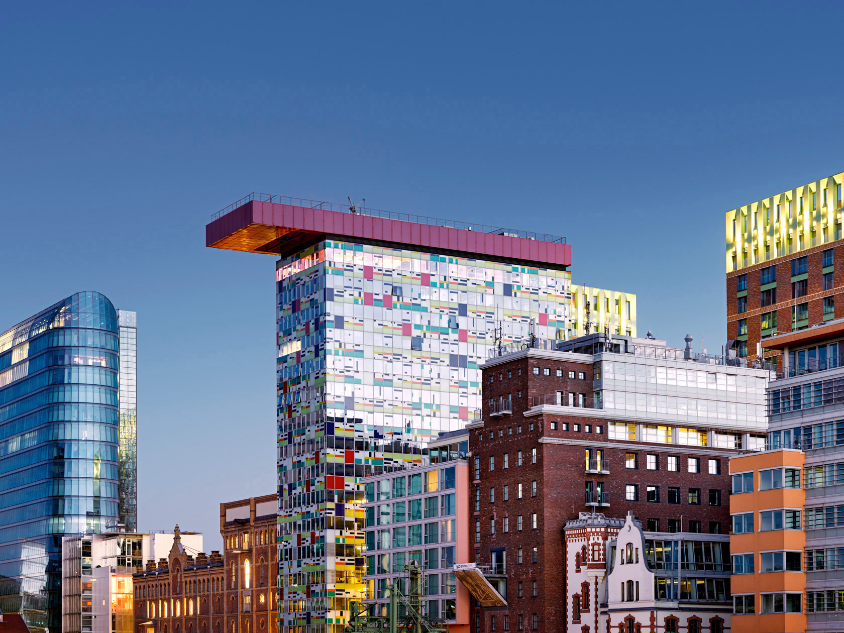 Herbert Böttcher zeigt die Architekturfotografie eines urbanen Stadtbilds in Düsseldorf mit farbiger Mosaikfassade, rotem Überhang, Glashochhaus links und historischen Backsteingebäuden bei klarem Tageslicht.