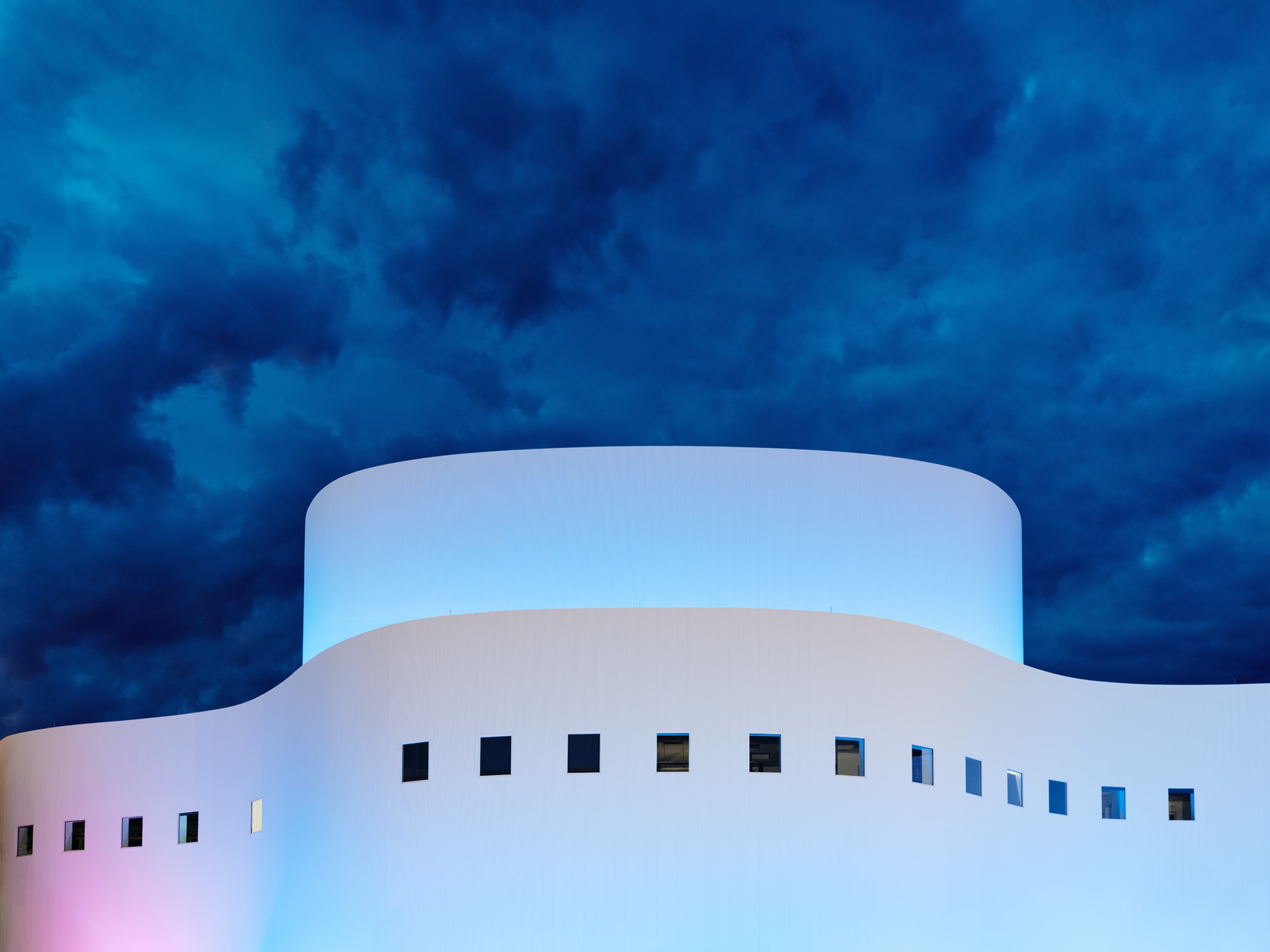 Die Fotografie von Herbert Böttcher zeigt das Düsseldorfer Schauspielhaus als ein modernes, geschwungenes Gebäude, das in blau‑rosa Licht getaucht ist und sich eindrucksvoll vor einem dramatischen Abendhimmel abhebt.