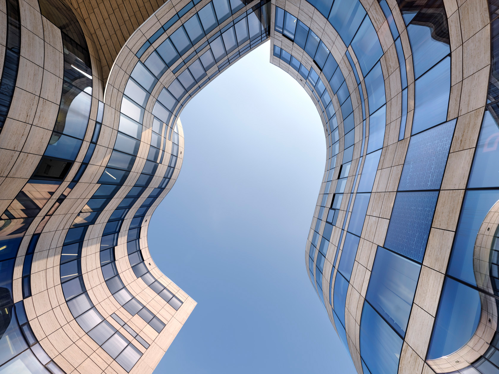 Die Fotografie Kö Bogen zeigt eine moderne geschwungene Fassade aus Glas und hellen Betonplatten, fotografiert von unten vor blauem Himmel.