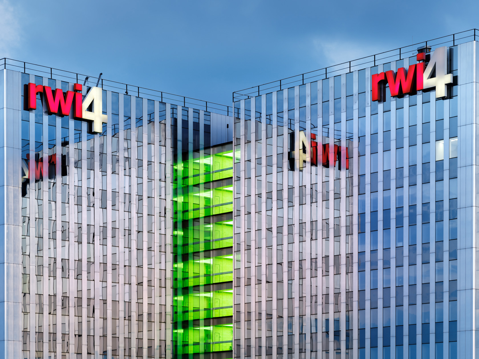 Die Fotografie des Düsseldorfer Fotografen Herbert Böttcher zeigt ein modernes Glas-Bürogebäude mit rotem RWI4-Logo und grün beleuchtetem Mitteltrakt und reflektierende Fassaden.