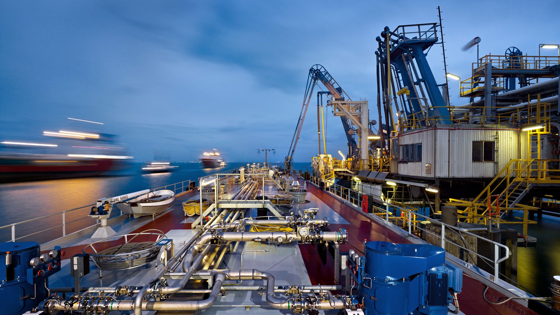 Industrieschiff im Hafen bei Dämmerung mit komplexen Rohrleitungen, Ventilen und Maschinen auf dem Deck; Kräne und weitere Schiffe im Hintergrund.
