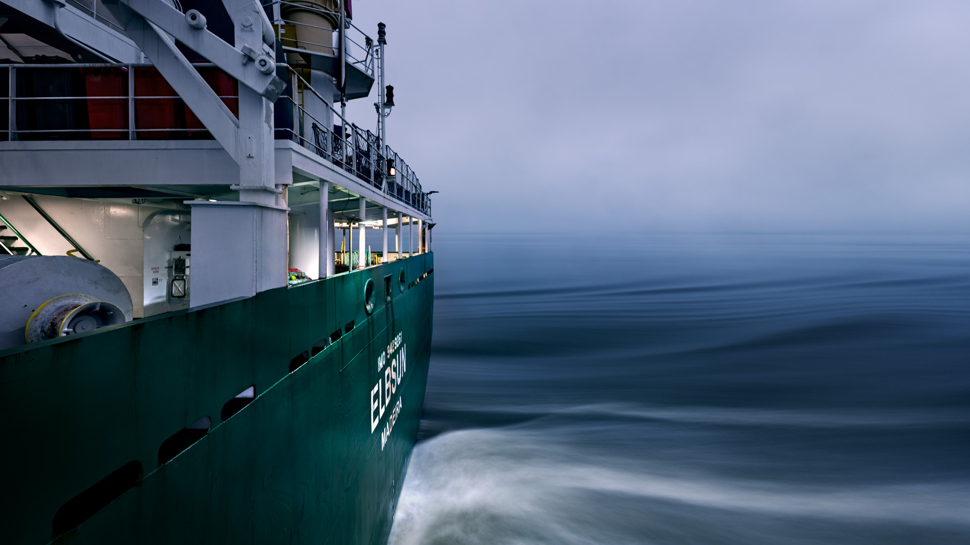 Sie sehen das Heck des Containerschiffs Ellbsun während der Fahrt in der Dämmerung. In seinem Langzeitprojekt Seamotion präsentiert der Fotograf Herbert Böttcher künstlerische Fotografien von Frachtschiffen auf den Weltmeeren. Die Bildreihe MS Elbsun entstand in Kooperation mit der Elbdeich Reederei, Drochtersen.