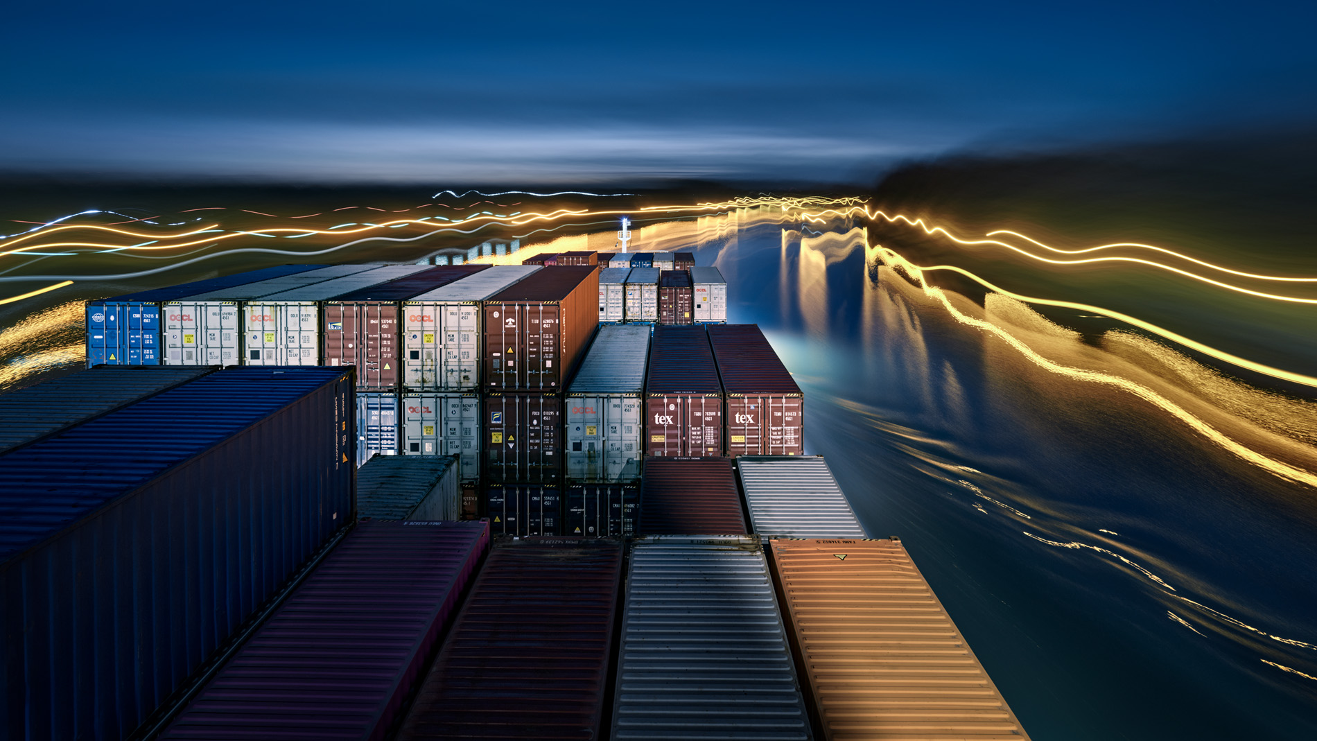 Blick vom Aufbau über dass Vorschiff des Containerschiffs Elbsun während der Fahrt auf dem Nord-Ostsee-Kanal in der Abenddämmerung. Es ist eine Langzeitbelichtung wodurch die Wasseroberfläche geglättet wirkt und Signale als gelbe Lichtspuren am Ufer sichtbar werden. In seinem Langzeitprojekt Seamotion präsentiert der Fotograf Herbert Böttcher künstlerische Fotografien von Frachtschiffen auf den Weltmeeren. Die Bildreihe MS Elbsun entstand in Kooperation mit der Elbdeich Reederei, Drochtersen.