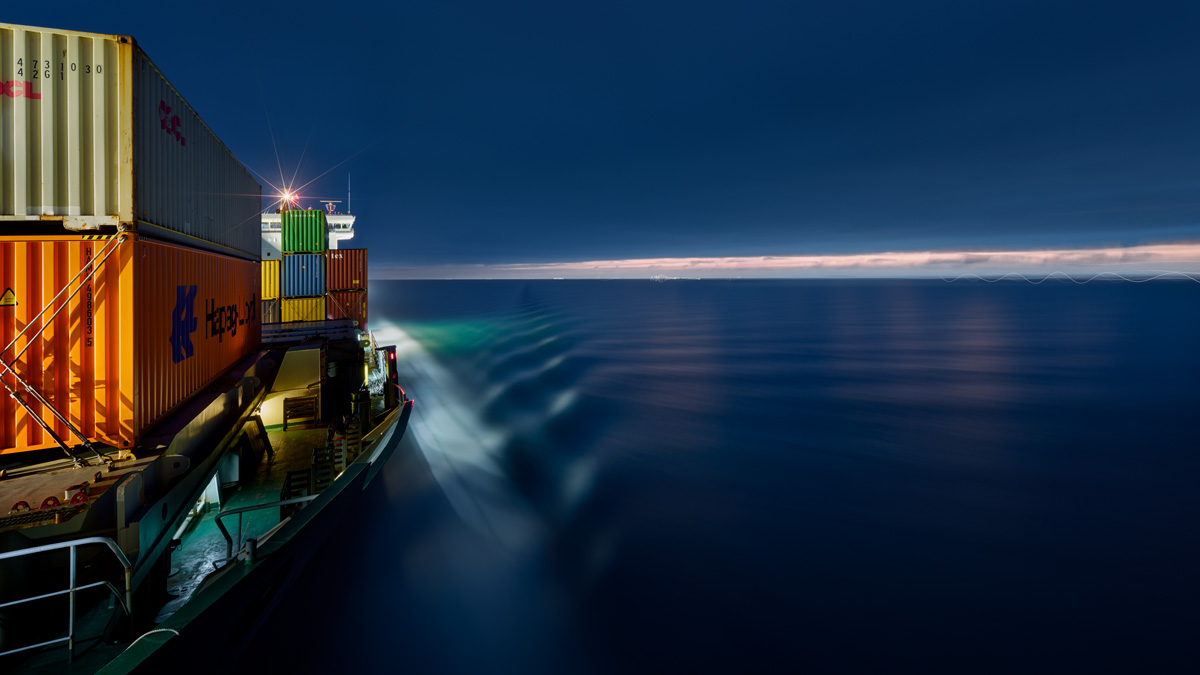 Sie sehen das mit Containern beladene beleuchtete Vorschiff des Containerschiffs Elbsun mit Blickrichtung zum Steuerhaus während der Fahrt in der blauen Dämmerung. Durch die Langzeitbelichtung wirkt die Oberfläche des Wassers geglättet.