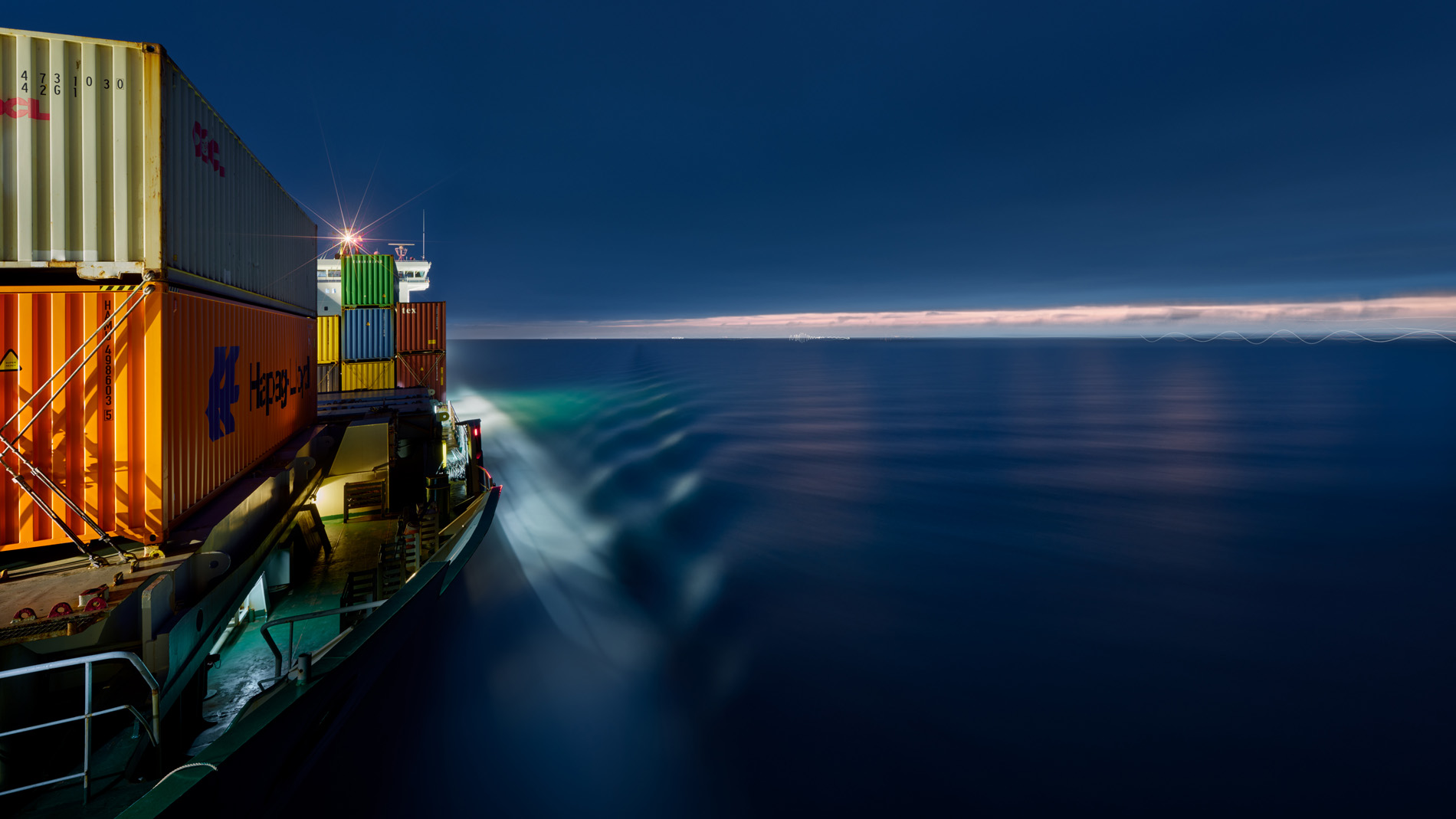Blick vom Bug über das beleuchtete Vorschiff und das Meer in Richtung des weißen Aufbaus des Containerschiffs Elbsun während der Fahrt in der Abdenddämmerung. In seinem Langzeitprojekt Seamotion präsentiert der Fotograf Herbert Böttcher künstlerische Fotografien von Frachtschiffen auf den Weltmeeren. Die Bildreihe MS Elbsun entstand in Kooperation mit der Elbdeich Reederei, Drochtersen.