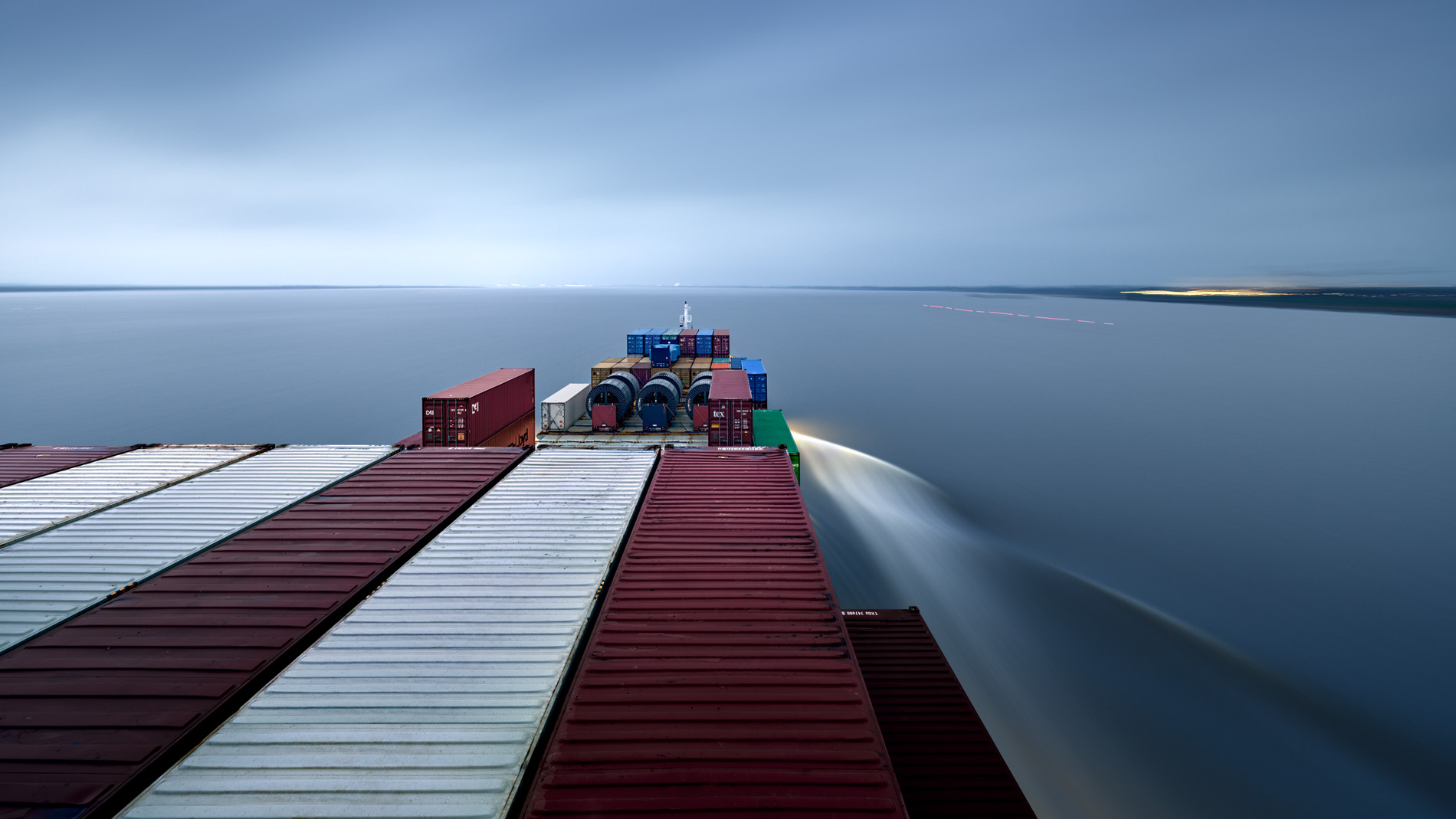 Blick vom Aufbau über dass Vorschiff des Containerschiffs Elbsun während der Fahrt auf der Elbe bei Brunsbüttel in der Abenddämmerung. Es ist eine Langzeitbelichtung wodurch die Wasseroberfläche geglättet wirkt, was die Stimmung des ruhigen Dahingleitens eindrucksvoll unterstützt. In seinem Langzeitprojekt Seamotion präsentiert der Fotograf Herbert Böttcher künstlerische Fotografien von Frachtschiffen auf den Weltmeeren. Die Bildreihe MS Elbsun entstand in Kooperation mit der Elbdeich Reederei, Drochtersen.