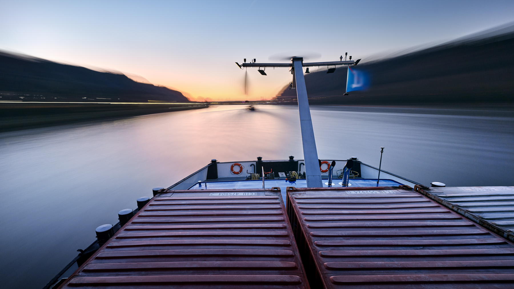 Blick auf Container und Bug eines fahrenden Containerschiffs bei Morgenlicht auf dem Oberrhein. Die aufgehende gelbe Sonne voraus und links und Rechts dunkle Hügel. Hügel und Wasser wirken durch die Bewegungsunschärfe geglättet. Waterway ist ein Projekt des Fotografen Herbert Böttcher aus Düsseldorf.