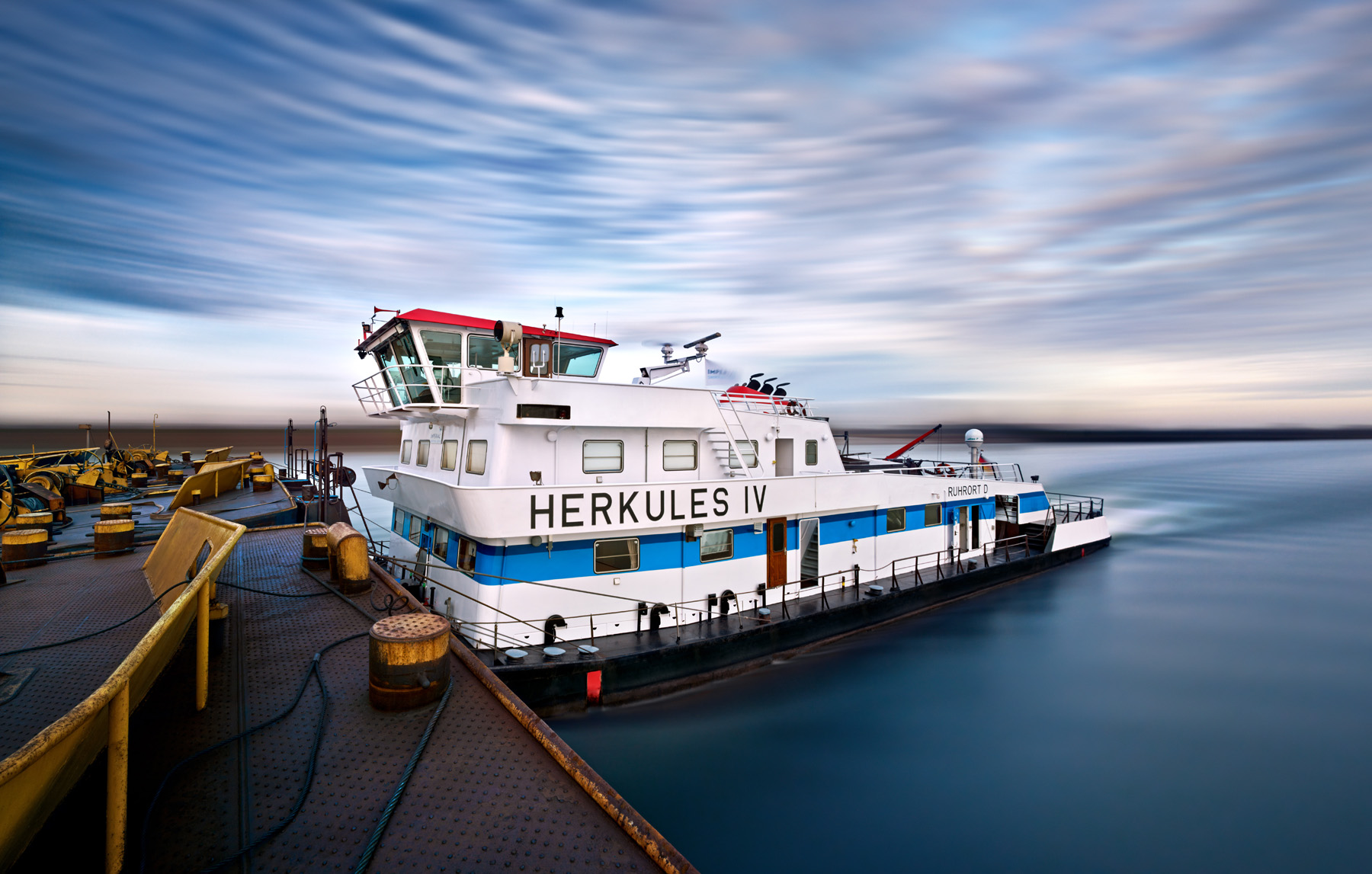 Die Fotografie zeigt das Schubschiff Herkules 9 während der Fahrt. Es ist eine Langzeitbelichtung; das Wasser und die ziehenden Wolken wirken dadurch glatter. Urheber ist der Fotograf Herbert Böttcher.