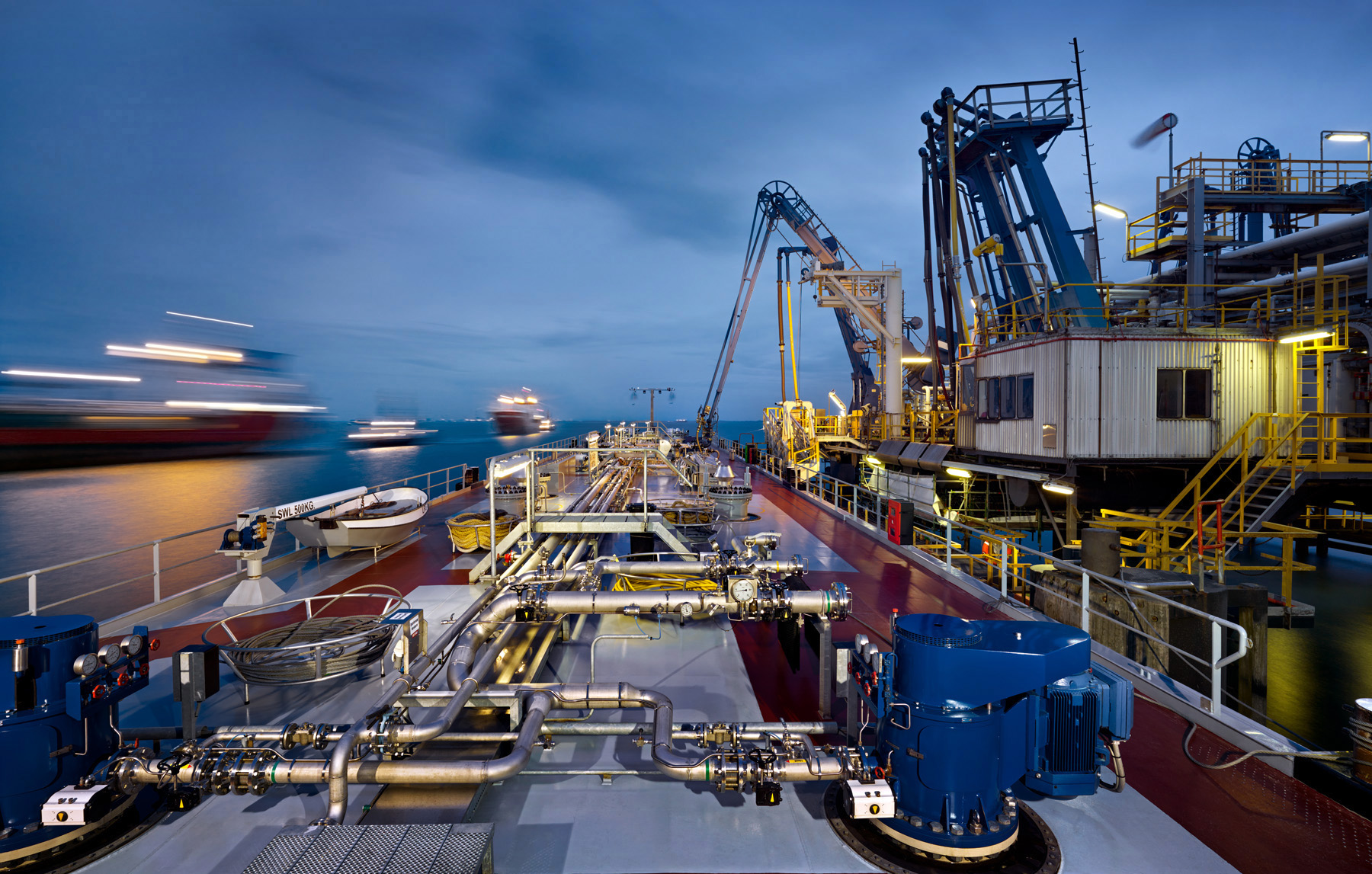 Die stimmungsvolle Fotografie zeigt ein Binnentankschiff an einem Verladeterminal bei blauem Abendlicht. Vorbeifahrende beleuchtete Schiffe erzeugen Lichtspuren. Fotograf Herbert Böttcher. Die Fotografie wird von HGK Shipping verwendet.